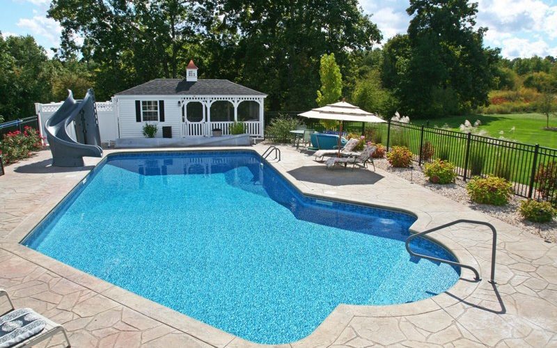 This is a photo of a Lazy L style custom inground swimming pool with a black fence, steps, water slide, and custom pool house.